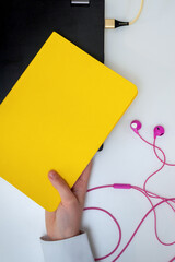 Flat lay with a yellow book for copy space and electronic devices - laptop and pink earphones on white background.