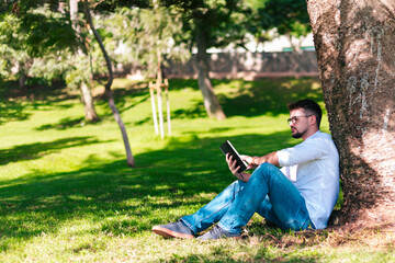 Obraz premium Young intelligent man wearing eyeglasses is reading book leaning on a tree in the city park