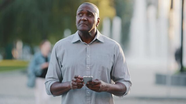 Confused adult mature african man outdoors in city park holding phone, looking around lost. Elderly black businessman trying to find building using electronic map of geolocation app in smartphone