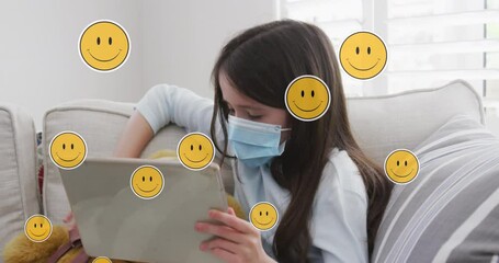 Animation of digital emoji icons over caucasian girl in face mask using tablet at home - Powered by Adobe