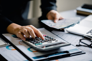 Close up of Businesswomen or Accountant using a calculator with counting dollar bills and analyzing business report graph, finance chart at the workplace, financial and investment concept.