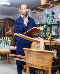 Portrait of positive professional man carpenter standing with book in furniture repair shop