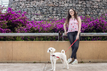 young white latina girl standing next to a garden of purple flowers with her white poodle dog.
