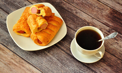 A cup of black tea and a plate with three sausages in dough on a wooden table.