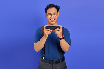 Cheerful asian young man playing video game on smartphone on purple background