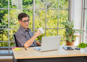 Asian executive senior businessman sitting on desk office with his office, the confident middle aged handsome man using laptop computer at workplace home office and drinking a cup of coffee