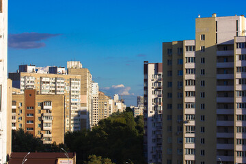 Fototapeta na wymiar Multistory buildings against a blue sky at sunny day. A city street stretching into a distance buried in green trees. Walls of high post soviet union buildings. Ugly architecture. View from a window.