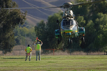 A medium-size utility helicopter is shown flying just above a field during the day, with a ground crew assisting with the landing.