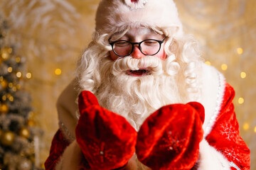 Cute santa claus in glasses on a wall background with a bright garland bokeh, hands in mittens...