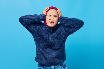 Portrait of angry young Asian woman covering her ears with hand on blue background