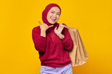 Beautiful Asian woman showing phone call gesture and holding shopping bags on yellow background