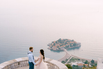 Fototapeta na wymiar The bride and groom are standing and holding hands on the observation deck overlooking the island of Sveti Stefan 