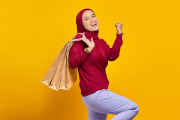 Excited and cheerful young Asian woman holding shopping bags on yellow background