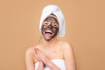 Mud facial mask, face clay mask spa. Beautiful laughing woman with cosmetic mud facial procedure, spa health concept. Skin care beauty treatment. Towel on head. Beige background, isolated.