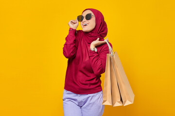 Obraz premium Cheerful young Asian woman wearing sunglasses and holding shopping bags on yellow background