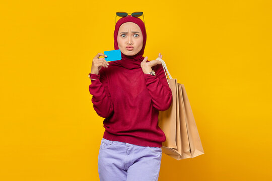 Shocked young Asian woman holding credit card and shopping bags on yellow background
