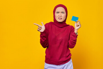 Shocked Asian woman holding a credit card while pointing finger at copy space on yellow background