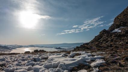 The frozen lake is surrounded by mountains. Snow on the ground. The sun is shining in the blue sky. Glare on the ice. Baikal