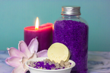 Obraz na płótnie Canvas Violet lavender sea salt for bath in the bottle, a burning candle and pink lotus flower in a background. A bar of soap or dry shampoo. Face and body care, spa treatments. Cosmetic products background.