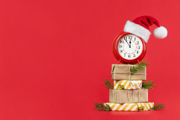 Red alarm clock in Santa Claus hat on wrapped gift boxes decorated fir branches and gold cones. Red...