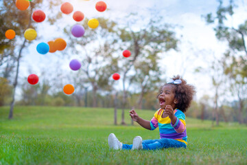 Cute little dark skinned girl very excited with colorful balls that was falling on her, sitting on...