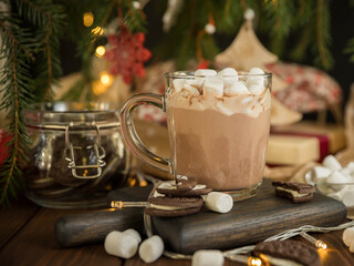 Hot chocolate with marshmallow  in a glass cup on a wooden table under the Christmas tree . Hot beverage and Christmas tree . Christmas greeting card.
