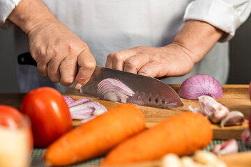 The hands of an adult woman cutting onion with a kitchen knife on a wooden board for lasagna preparation.