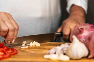 An unrecognizable adult woman using both hands on a kitchen knife to chop a garlic on a cutting board for lasagna preparation.