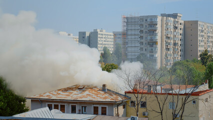 View of firefighters using water to extinguish fire from burning house. Smoke and flames created by fire from building roof spreading dangerous smog and fumes in city neighbourhood.