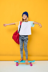 Charming little blonde kid girl rides a skateboard with a red backpack isolated background