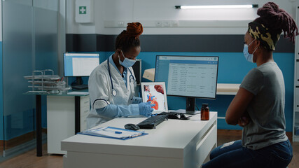 Medic with face mask pointing at tablet with heart figure for examination with patient in cabinet. Doctor explaining cardiovascular diagnosis to woman while protecting against coronavirus