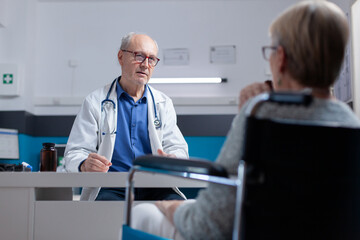 Close up of doctor and woman with disability meeting at checkup visit in cabinet. General practitioner doing consultation and examination for patient sitting in wheelchair, receiving help.