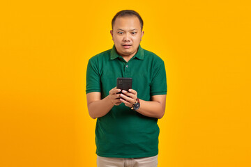 Shocked handsome young Asian man looks at smartphone screen isolated on yellow background