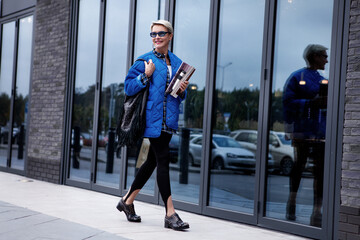 Fashionable woman with short hair walking street dressed in stylish casual outfit, blue jacket and sunglasses, black leather handbag and boots, leggings with straps. Trend spring or autumn - obrazy, fototapety, plakaty