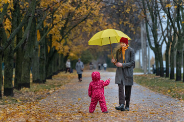 Mom walks with small child in an autumn park under a yellow umbrella