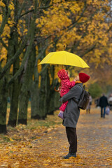 Obraz premium Mother holds a small child in her arms while standing under a yellow umbrella in an autumn park against a background of yellow foliage. Vertical frame