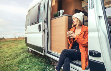 Woman drinking coffee sitting at the door of a camper van - obrazy, fototapety, plakaty