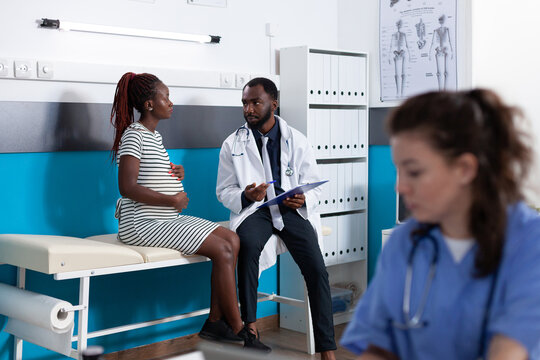 African American Woman And Doctor Talking About Pregnancy At Medical Appointment In Office. Patient Expecting Child And Having Discussion With Physician To Check Healthcare. Pregnant Adult