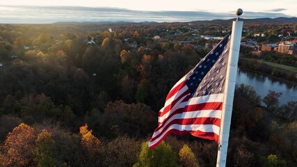 Cinematic aerial of American flag waving in breeze during autumn golden hour sunrise light. Colorful foliage, sunlight. America the Beautiful, Proud to be an America. Town reflects in river. - Powered by Adobe