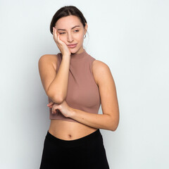 Fototapeta na wymiar Cute young woman standing in short turtleneck, holding palm on cheek and leaning face on it, being bored and tired of uninteresting talks, feeling indifferent to what happening over gray wall