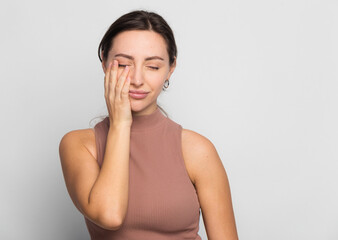 Cute young woman standing in short turtleneck, holding palm on cheek and leaning face on it, being bored and tired of uninteresting talks, feeling indifferent to what happening over gray wall