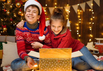 Portrait of a boy and girl in New Year decoration. They open a box and have fun. Holiday lights, gifts and a Christmas tree decorated with toys.
