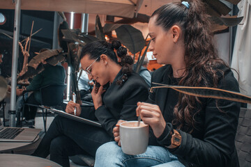 Compañeras de trabajo con seriedad llamando por telefono movil y escribiendo planteamientos junto su ordenador laptop en una cafeteria de su ciudad tomando café por la mañana para terminar su trabajo