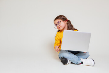 Cute little girl with glasses having video lesson with laptop on gray background, studying with...