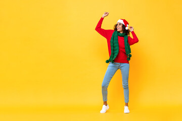 Fototapeta na wymiar Full length portrait of fun happy Afircan American woman in Christmas attire dancing on isolated yellow studio background with copy space