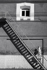 A young woman in a raincoat and dark glasses, with a suitcase in her hands, climbs the fire escape against the background of a brick wall. Retro style black and white outdoor shot