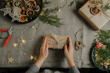 Female hands holds Christmas present wrapped in kraft paper. DIY Christmas packaging for holiday...
