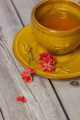 Obraz na płótnie Canvas Ceramic yellow cup with black tea stands on saucer on grey wooden table. Spindle shrub, Euonymus europaeus, Pfaffenhutchen red flower petals. Petal in a form of heart. Flat lay still life composition.