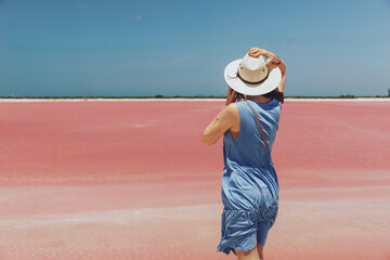 A young woman in a hat and blue dress stands against the backdrop of a salty pink lake in Mexico,...