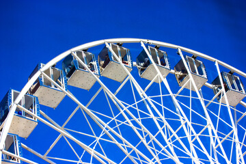 A white Ferris wheel with transparent booths on blue sky background. City entertainment. Luna park games, fun for kids, carrousel. High construction view from below. An attraction for gamblers. 